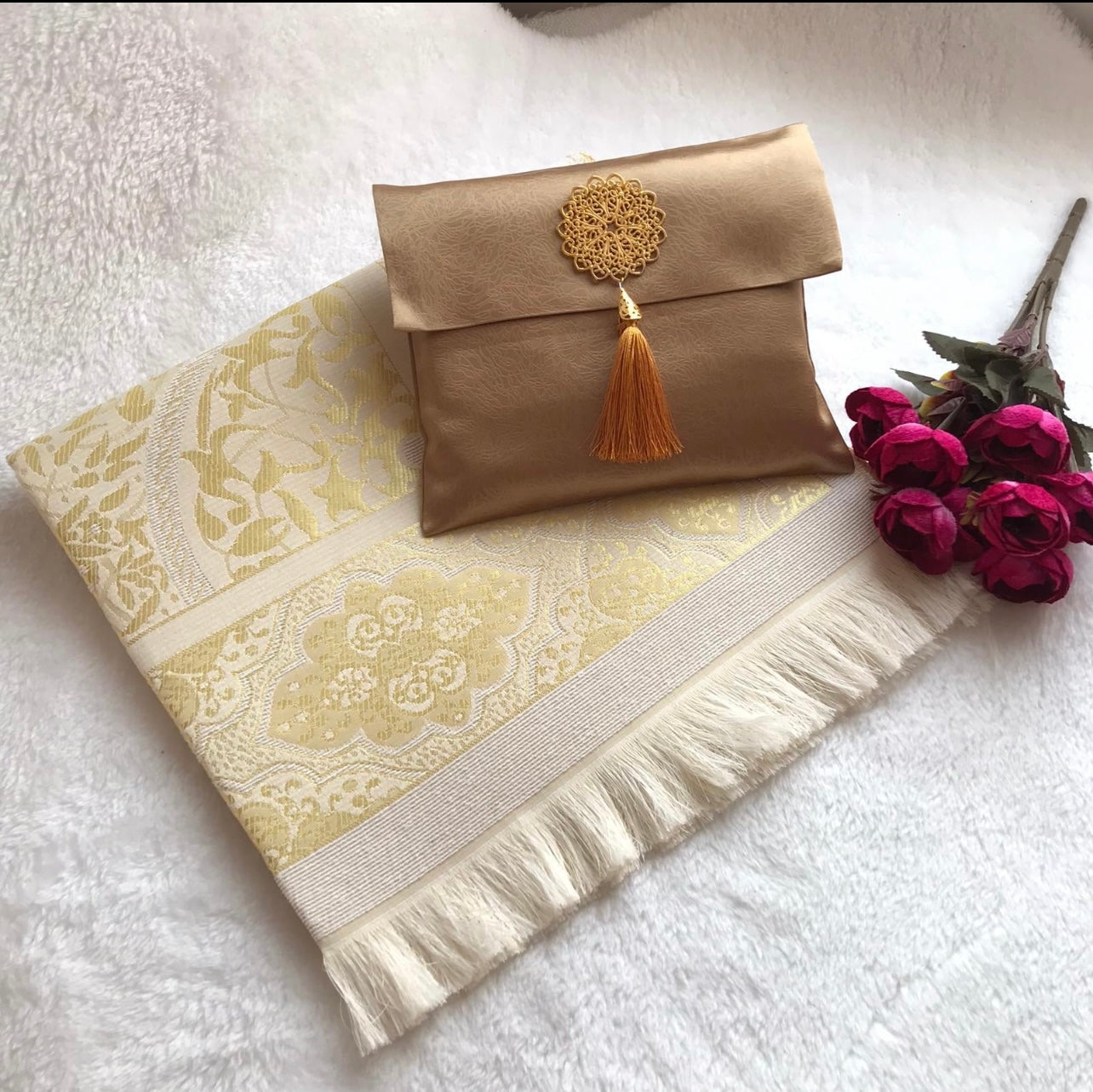 A gold-colored rectangular prayer rug with a floral pattern, displayed alongside a beige-colored handbag with a tassel, and a white tassel on a white surface.