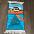 A colorful prayer rug featuring an illustration of a cityscape with a boat on water, fringed with a tassel on the bottom.