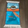 A colorful prayer rug featuring an illustration of a cityscape with a boat on water, fringed with a tassel on the bottom.