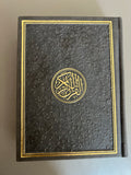 A black leather embossed Quran with gold trim, featuring the Arabic text and decorative design on the cover.