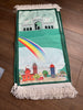 A green prayer rug with a colorful cityscape design, featuring a rainbow, buildings, and a mosque.