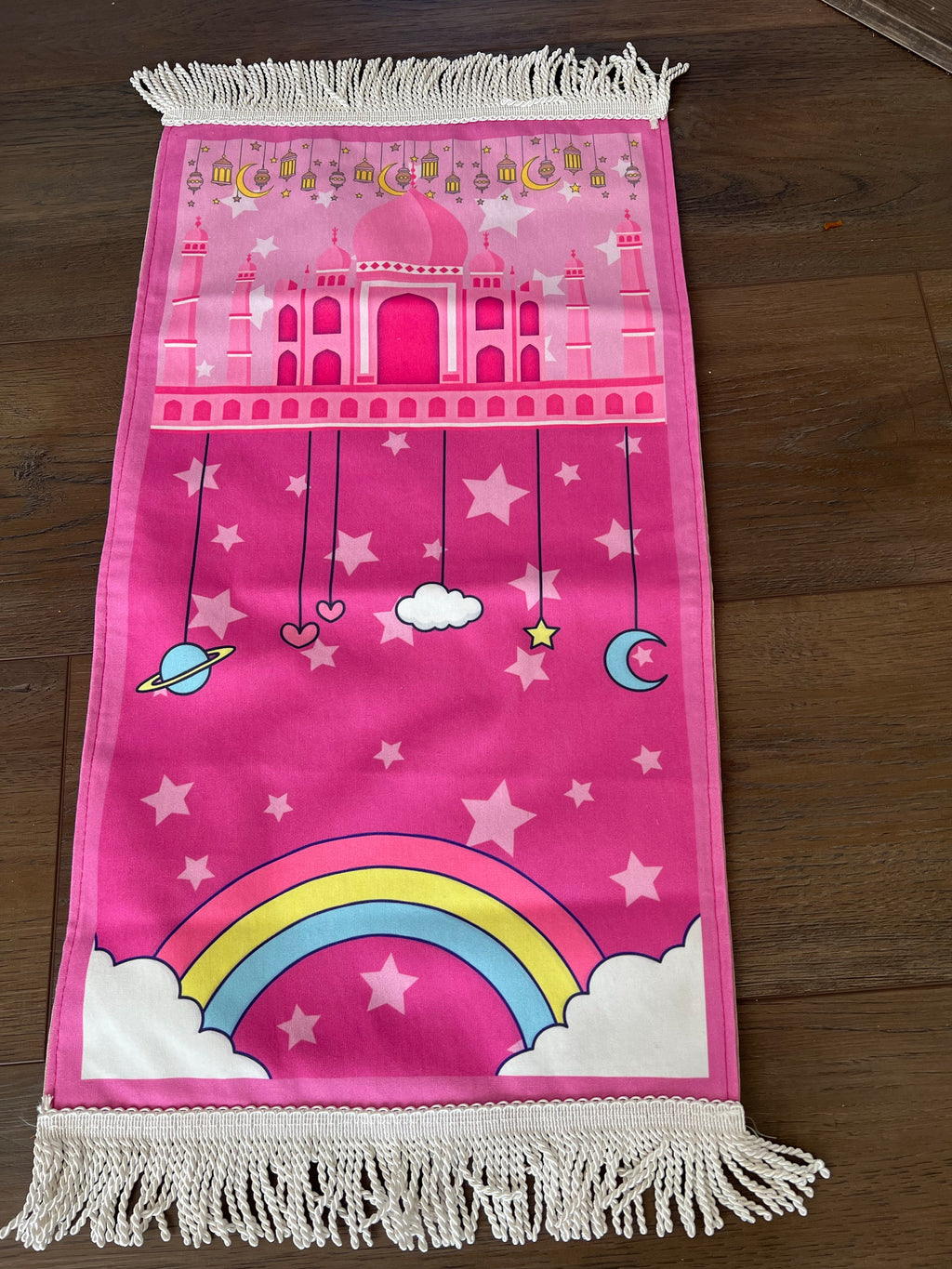 A pink prayer rug featuring a space-themed design with stars, planets, and a mosque, laid out on a wooden floor.