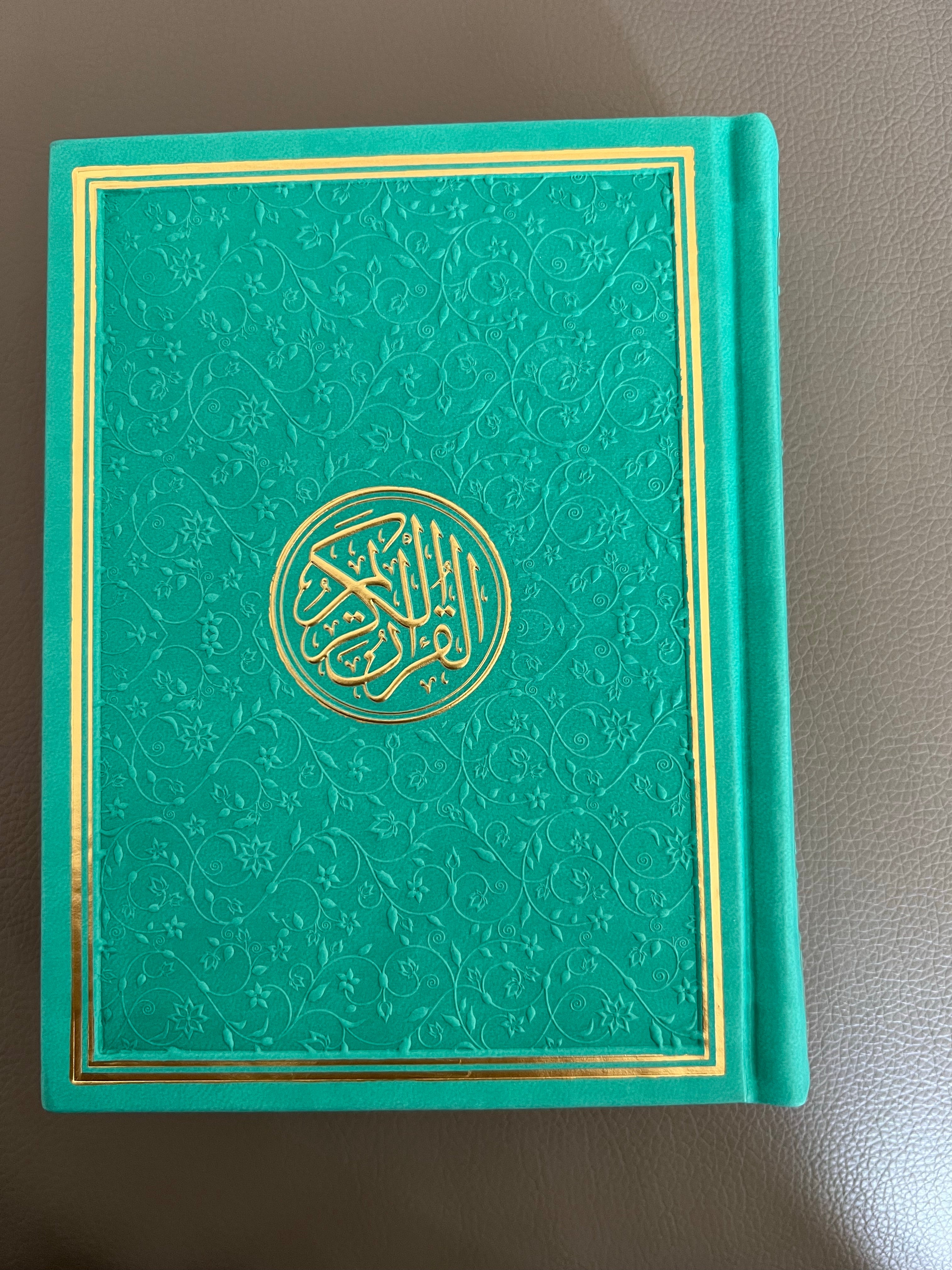 A teal green Quran with gold trim on the cover, featuring Arabic text and decorative gold patterns.