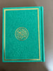 A teal green Quran with gold trim on the cover, featuring Arabic text and decorative gold patterns.