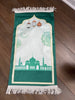 A green prayer rug featuring a white and green geometric pattern with an illustration of a mosque and stars.