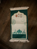 a prayer mat with a dark green border, a mosque at the border, drawings of flowers, and brown and gold colored lamps above