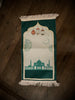 a prayer mat with a dark green border, a mosque at the border, drawings of flowers, and brown and gold colored lamps above