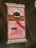 A pink and purple prayer rug with a cartoonish design featuring a mosque, stairs, and a boat.